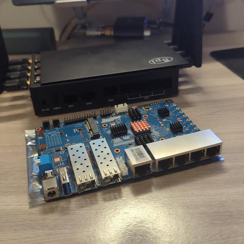 Una placa de circuito impreso azul, Banana pi BPI R3, se encuentra sobre una mesa de madera. La placa tiene varios conectores, incluyendo un conector de alimentación, múltiples puertos Ethernet y dos ranuras para tarjetas SFP. Junto a ella, se observa una caja negra rectangular, que podría ser un router o un módem, con varios puertos en su parte trasera. Ambos dispositivos parecen ser parte de un equipo de red.