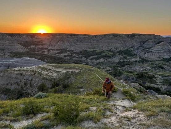 A man hikes up a hill in the North Dakota badlands while the sun rises on the horizon in the background.