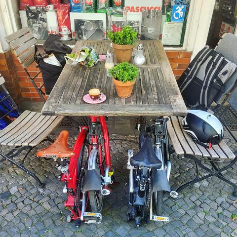 Ein Holztisch mit vier Biergarten-Holzstühlen vor einem Ladengeschaft mit Espressopackungen im Schaufenster. Auf dem Tisch 2 Blumentöpfe,  2 Zuckerspender, 1 Espresso. Unter dem Tisch in Bildmitte unten sind nebeneinander eingereiht und eingefaltet 2 Bromptons. Das linke ein roter Rahmen mit Leder-Vintage-Sattel, das rechte ein schwarzer Rahmen mit sportlichem schwarzen Sattel. Auf dem rechten Stuhl ist ein schwarz-weißer Fahrradhelm abgelegt. Auf den hinteren beiden Stühlen liegen Taschen und Rucksack.