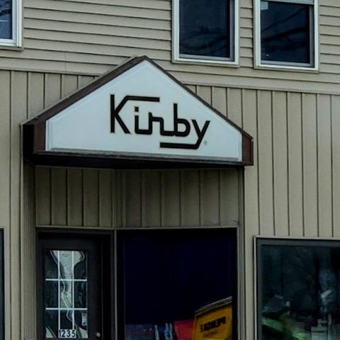 A triangular sign above a building entrance that reads "Kirby". The background of the sign is white and the logo is a black, squarish cursive.