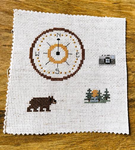 A cross stitched design with a large compass rose in the upper left of an oatmeal canvass, with smaller designs around the lower to middle left of the compass depicting a brown bear, a sunrise between some pine trees and a tiny camera.