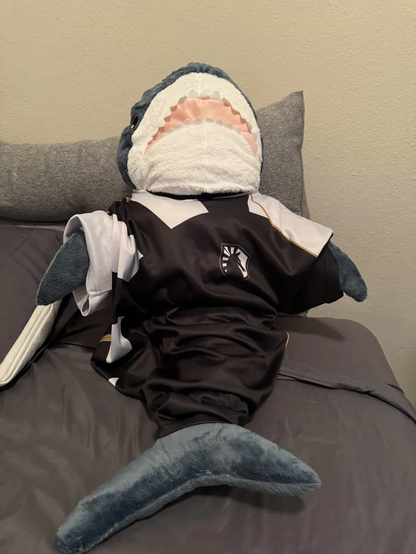 A BLÅHAJ shark plushie dressed in what appears to be a Team Liquid esports jersey (black with white accents and team logo). The shark is positioned on a bed or couch with gray bedding, propped up against a pillow with its open mouth and distinctive teeth visible. The plushie is arranged to look like a fan watching or cheering for Team Liquid at the CS2 tournament IEM Melbourne. The blue shark's tail is visible at the bottom of the image, and the entire setup creates a playful scene of the IKEA shark as an enthusiastic esports supporter.