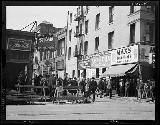 The image is a black-and-white photograph depicting an urban street scene from what appears to be the early 20th century. The setting includes buildings with signage indicating various businesses, such as "STEAM," "Coca-Cola," and shops like Max's that advertise items for men including gloves.

There are several individuals present on the sidewalk; most of them seem engaged in conversation or looking around, possibly waiting to cross a street indicated by signs reading 'Danger Street Closed' and other warnings. The attire suggests it is from an earlier time period, likely during the Great Depression era considering the context provided for this photo being part of Dorothea Lange's work.

The overall atmosphere conveys a sense of daily life in a working-class neighborhood with people going about their business despite challenges implied by economic conditions at that time. The photograph captures a slice-of-life moment and is likely intended to document social issues or the impact on everyday citizens during this period, as suggested by its inclusion among works associated with Dorothea Lange's photojournalistic efforts in San Francisco.

This image can provide insights into urban life and socioeconomic conditions of America around that time.