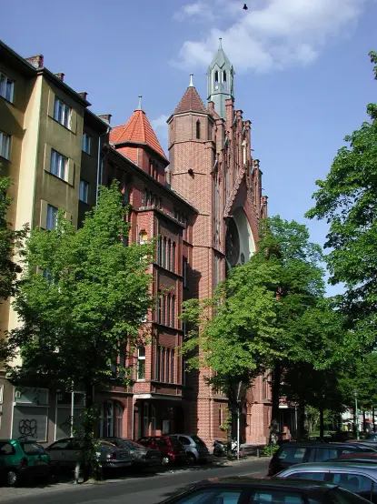 The image depicts a street scene featuring a combination of buildings, including a prominent brick structure with a tower, possibly a church or historical building. The architecture appears to be influenced by the Gothic style, with pointed arches and decorative elements. There are trees lining the street, and the scene captures a clear blue sky with some clouds. Vehicles are parked along the street, suggesting an urban environment.

Image Credits: Wikimedia / Angela Monika Arnold / CC BY-SA 2.0 de
