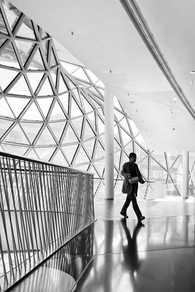Black and white. Portrait format. A shopping center in Frankfurt with striking steel/glass architecture.  Interior shot. A passer-by in the backlight of the glass façade appears as a faceless black silhouette reflected in the smooth floor covering. There is a strong contrast between the bright white interior and the silhouette.