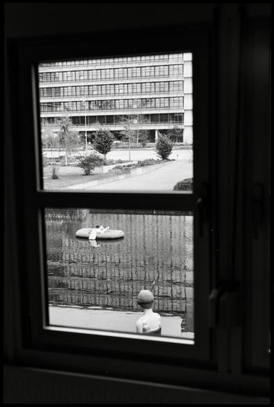 Black and white film photograph. View out of a window with two panes. In the upper part of the window, a building with a sidewalk is visible. In the lower part, a swimming pool with installed figures depicting the rescue of a drowning man.