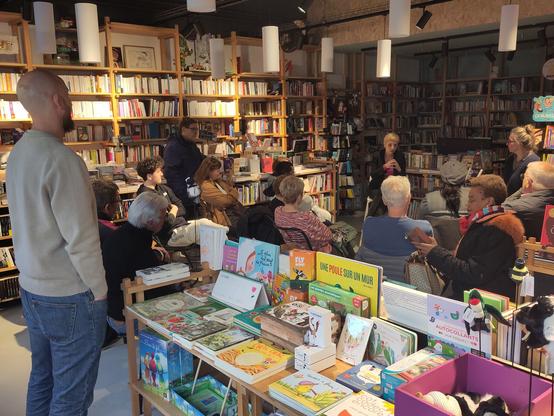plusieurs personnes debout et assises écoutent un débat dans une librairie. au premier plan et en arrière-plan, des rayonnages de livres.