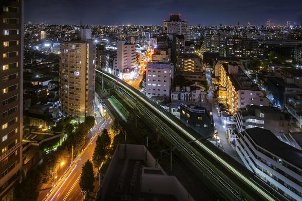 View on Tokyo at night from a skyscrapper building, using a slow shutter speed. A train is crossing the image in diagonal creating a green glow on the black tracks. Buildings and streets around it are bathed in orange or blue lighting depending on the types of lamps used.