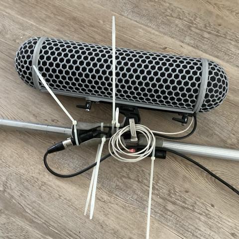 A Rode blimp windshield fastened to a aluminium stick by it’s pistol grip, using a whole bunch zip ties. There are white and black cables running from the blimp, neatly zip tied to aluminium stick. The excess of white cable has been rolled up and affixed under the pistol grip lock lever.