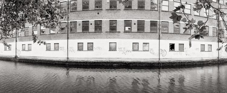 Captured in a stark black and white, a curved brick building stands firmly at the water's edge. The lower portion of the structure is painted a lighter shade and bears faint graffiti, contrasting with the darker brick above, punctuated by numerous rectangular windows. The still water reflects the building's facade, creating a mirrored image with subtle ripples disturbing the surface. Framing the scene are leafy branches in the foreground, adding a touch of organic texture to the otherwise architectural focus. The overall mood is one of quiet urban stillness.