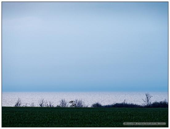 Streng dreigeteiltes Bild; das untere Bilddrittel teilen sich kurz vor Sonnenuntergang ein in dunklem, mattem Grün daliegender Acker mit den Silhouetten einzelner Sträucher am Horizont und darüber die im Gegenlicht silbrig glänzende Ostsee, darüber ein bleigrauer, konturloser Himmel.