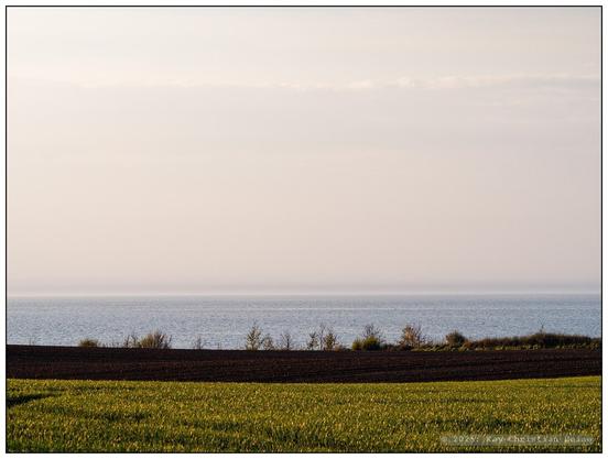 Wie oben, aber früher fotografiert: Der Himmel ist noch blassblau mit leichter Wolkenstruktur, die Ostsee schimmert bläulich und das Ackerland im Vordergrund ist am unteren Bildrand grün von jungem Getreide, darüber das Dunkelbraun eines frisch bearbeiteten Ackers.