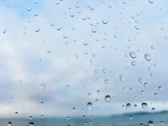 Durch Licht und Schatten klar abzeichnende Wassertropfen verteilt auf einer Fensterglasfläche, die den Blick ins graue weichgezeichneten Wolkenhintergund mit dunkelgraublauem unscharfen Horizont in der Ferne freigibt.