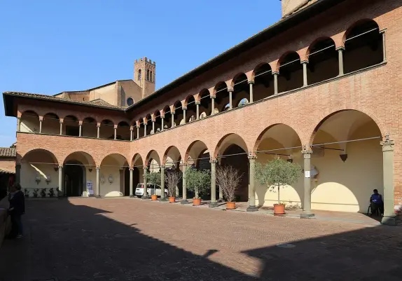 The image features an architectural exterior of a building with a courtyard. It showcases a series of arches along the ground level, with columns supporting the upper structure. The upper level has windows with decorative features. In the background, there's a tower visible, and the scene appears to be set in a sunny environment with clear blue skies. The courtyard includes potted plants, and there are a few people visible, contributing to the lively atmosphere.

Image Credits: Wikimedia / Sailko / CC BY 3.0