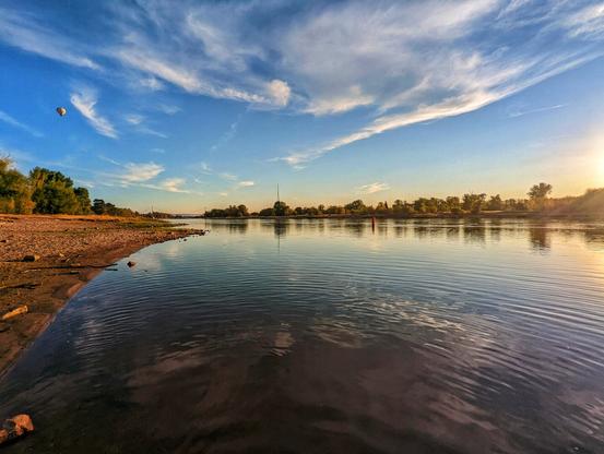 A wide shot captures a serene river scene at dusk. The sky above is a blend of clear blue and wispy white clouds, with the sun, a bright orb, positioned on the right horizon, casting a golden glow across the water. The river itself is calm, reflecting the sky and the line of green trees that borders its far bank. The near bank is a mix of sandy and rocky terrain, leading down to the water's edge. A lone hot air balloon floats gently in the upper left of the frame, adding a touch of whimsy to the tranquil landscape.