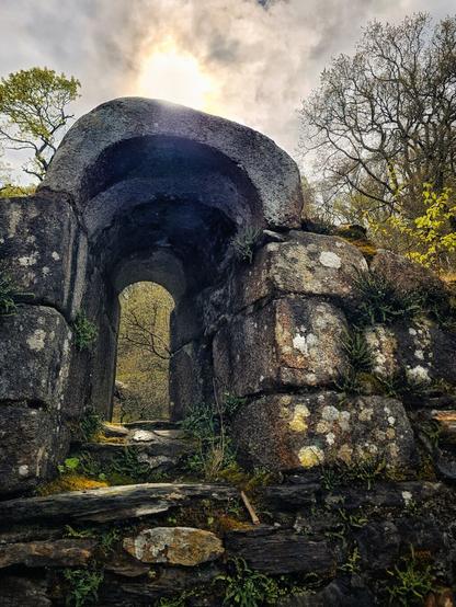 A low-angle shot of an old stone archway in a forest, with sunlight filtering through the opening.