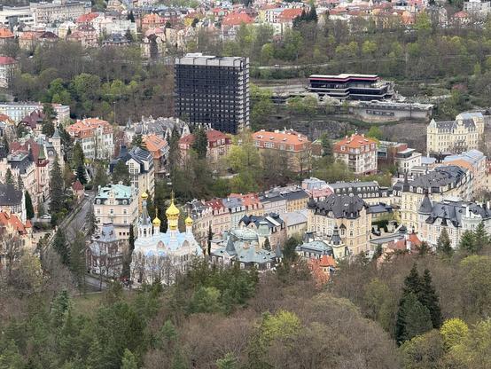 Das Bild zeigt eine Luftaufnahme einer Stadtlandschaft, wahrscheinlich eines Kurortes. Im Vordergrund ist ein Waldstück zu sehen, dahinter reihen sich zahlreiche Gebäude aneinander, überwiegend im Stil von Villen und Hotels, die eng beieinander stehen.  Eine blau-weiße Kirche mit goldenen Kuppeln ist deutlich im mittleren Bereich des Bildes sichtbar.  Ein moderner, hoher Gebäudes steht etwas abseits der anderen Bauten. Die Architektur der Häuser deutet auf eine geschichtsträchtige, wohlhabende Gegend hin. Das Gesamtbild wirkt ruhig und vermittelt einen Eindruck von geordneter Architektur inmitten von Natur.