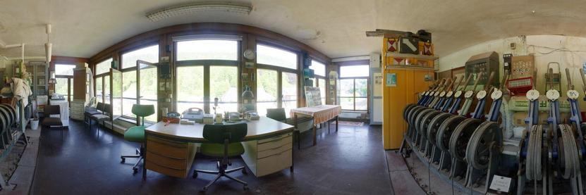 A panoramic view inside a signal box. The building appears to be curved as the viewer is offered a 180 degree angle of view. In the centre is a desk and a few office chairs. Opposite the viewer is a wide window front. On either side of the viewer, batteries of large levers pull you along, which were used to adjust the signals and points