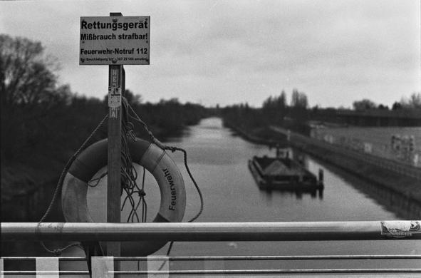 A black and white photograph of a live preserver on a bridge over a canal. The canal including a ship on it is out of focus. At the bottom one can see the effect of a light leak during processing of the film.