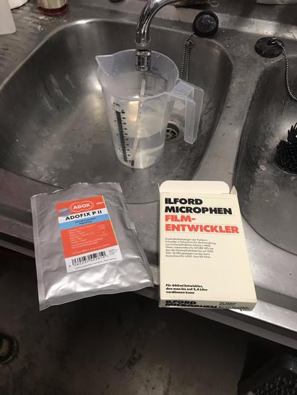 A color photograph of a sink with two packages of dry photo chemicals on it: Ilford Microphen and Adox Adofix P II.