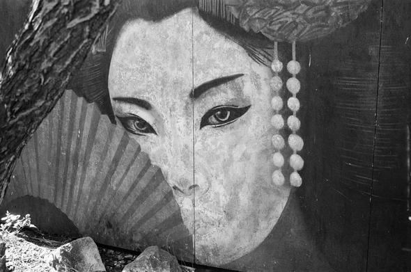 This black-and-white photograph captures a close-up of a large, weathered mural painted on a textured wall. The mural depicts the face of a woman with striking, almond-shaped eyes and delicate features. She wears a string of pearls draped along her cheek, adding a touch of elegance. To the left of her face, a partially visible fan adds an element of intrigue. The mural shows signs of age and wear, with visible cracks and faded areas, enhancing its rustic charm. The overall mood is nostalgic and artistic, highlighting the beauty of the subject amidst the passage of time.