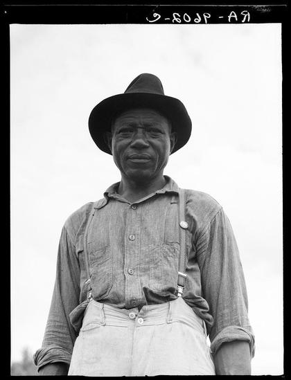 This black and white photograph features an older Black man standing outdoors. He is wearing a dark hat, a light-colored button-up shirt with rolled up sleeves, suspenders holding up striped trousers, and appears to be smiling slightly at the camera. His expression seems contemplative or content.

The background of the image is overexposed, suggesting it may have been taken on an unusually bright day or in high-contrast conditions which has caused some detail loss. The text "3-SOCP-A9" handwritten across the top indicates that this might be a photograph from a collection documenting social issues during the time.

The man's attire suggests he could be working class, possibly engaged in agricultural work given his practical clothing and setting. His union button on his shirt signifies membership or support for labor unions which were particularly active among American workers during certain historical periods of economic change and development.