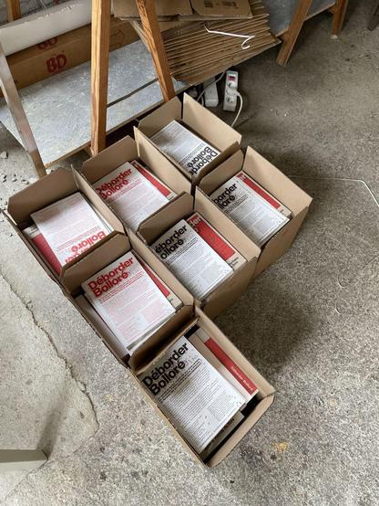 Photographie couleur de cartons ouverts, posés au sol, contenants des exemplaires du livre mentionné dans le toot.