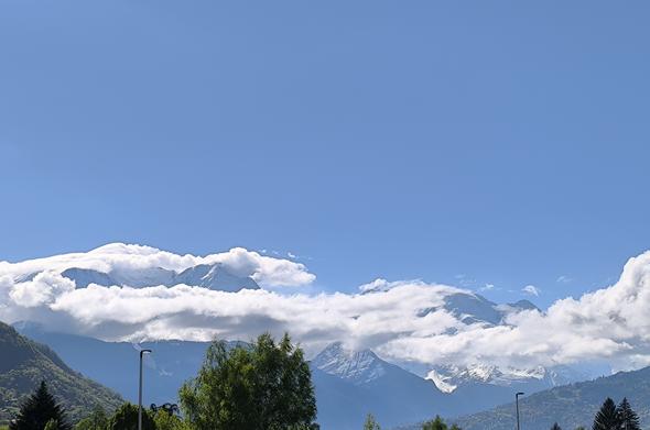 Vue sur le massif du Mont-Blanc. Le ciel est uniformément bleu et des nuages blancs s'accrochent autour des montagnes, comme des bandes de coton.