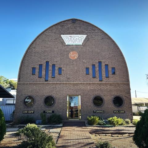 A brick building with a curved roof, designed to look like an old mid-century radio set. Under the four round windows (two on either side of the central door) are signs “on-off”, “tone”, “tuning” and “volume”. In the middle up top is a panel that shows some radio station codes. It’s a clear blue sky above.