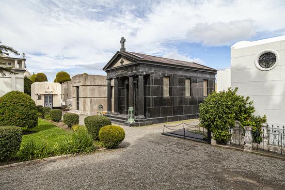 It's a sunny day with patchy, thin sheets of cloud in the sky. We're in a grand public cemetery comprising avenues of gravel between beautiful mausoleums of stone and marble. Directly in front of us there is one in black stone blocks, reminiscent of ancient Greek or Roman buildings, with a shallow, sloped roof and a portico with four fluted columns in front of the door. Nearer to us on the right there is one in white blocks with a round window set high up and a small railed-off forecourt around it with a few pretty bushes. More mausoleums of varying designs are to the left across a small section of lawn and topiary, each one no more than a few metres tall.
