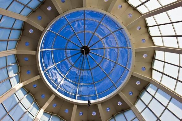 A glass dome photographed from the inside and from below. The dome fills the entire photo and consists of iron and glass. The dome is constructed in two layers. The lower, conical ring contains frosted glass panes and the upper, spherical ring clear glass, which is now brightly colored blue by the sky. Thirteen iron rings run from the center outwards and three round rings divide these into separate clear glass panes. Around the upper glass dome a wide iron scalloped edge and below that the frosted glass panes. Again divided by iron arches and rings into seperate panes.