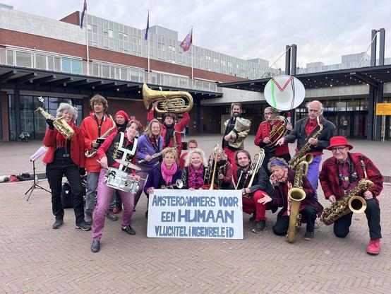 Straatorkest met al hun instrumenten en een bord waar op staat: "Amsterdammers voor een humaan vluchtelingenbeleid"