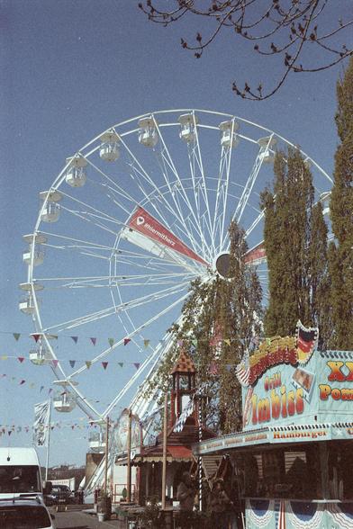 Das Riesenrad am Beuler Rheinufer, Blickrichtung Bonn. Im Vordergrund ein Imbissstand, dazwischen Bäume.