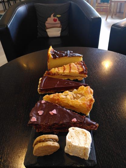Auf einem dunkelbraunen, runden Holztisch steht eine längliche Kuchenplatte aus Schiefer. Darauf befinden sich fünf verschiedene Kuchenstücke sowie ein Plätzchen und ein Marshmallow.
Von oben beginnend thronen folgende Stücke auf der Platte:
- Flan pâtissier (ein recht hohes Stück Kuchen, bestehend aus einem dünnen Mürbeteigboden  und -rand sowie einer etwa 5 cm hohen Puddingschicht, die mit einer gut gebräunten Haut abschließt)
- Caramel Shortbread (ein Mürbeteiggebäck in Kuchenform; zwei Schichten buttriger Mürbeteig, gefüllt mit meinem Karamell, verziert mit Streifen desselben Karamells)
- Schokotarte mit in Rum flambierten Bananen (auf einem Mürbeteigboden befinden sich die Bananenscheiben, darüber befindet sich eine etwa 1 cm dicke Schicht Schokoladenganache)
- Birnen-Mandel-Tarte (mit Vanille und Zitrone pochierte Birnenhälften betten sich in eine Mandelcrème auf buttrigem Mürbetwigboden; verziert mit Mandelblättchen)
- Sacher Shortbread (zwei Schichten Schokoladenmürbeteig, gefüllt mit Aprikosenkonfitüre, verziert mit Edelbitterschokolade sowie kleinen rosafarbenen Schokoladenherzen)
- links zwei Mandelplätzchen, die mit einem Pistazienkaramell zusammengesetzt sind und rechts ein Marshmallow mit Karamell.
Hinter dem Tisch steht ein schwarzer Ledersessel mit einem kleinen Kissen im Rückenbereich. Das Kissen trägt einen Aufnäher in Form eines Cupcakes mit einer Kirschverzierung.