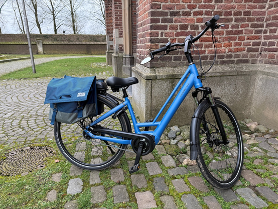 Baumattes Damenfahrrad der Marke Velodeville mit Nabenschaltung. Dicker, weicher Sattel an einer gefederten Sattelstütze. Am Gepäckträger sind Satteltaschen angebracht, die etwas dunkelblauer sind. Es handelt sich um ein Pedelec, bei dem der Akku auf dem Gepäckträger angebracht ist.