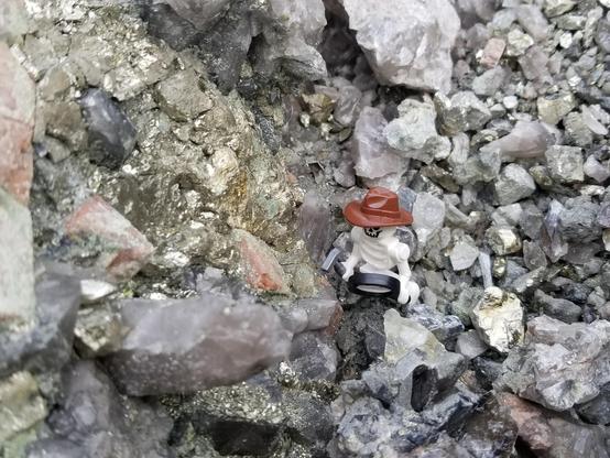 Lego skelleton with hat, hammer, and magnifying glasses standing in an agggregate of minerals. Pyrite and chalcopyrite are shiny silver-gold, quartz is greyish-blue and feldspar is orange.