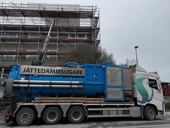 En stor tankbil parkerad utanför flerfamiljshus under renovering. En skylt står längst bak på tanken: “JÄTTEDAMMSUGARE”