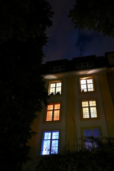 The image shows a building at night, partially obscured by trees. The windows are illuminated in various colors: one window glows orange, another blue, and a third emits a warm white light. The sky is dark with some clouds, creating a moody atmosphere.