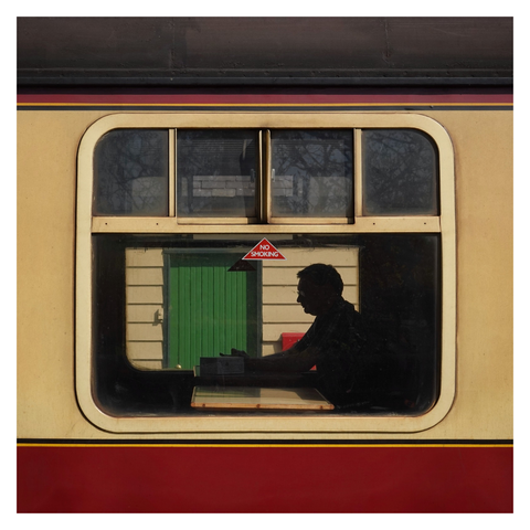 This is a square format colour image taken through the window of an old fashioned train carriage. A man is sat at a table on the far side of the carriage and is in silhouette. Taken in Pickering, Yorkshire, with a Sony RX10.