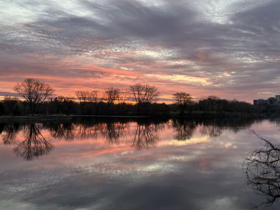 Streaks of orange pink skies with grey clouds full of texture reflect on the calm river with a treeline/cityscape silhouetted mid frame