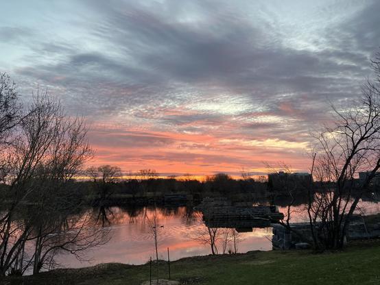 Streaks of orange pink skies with grey clouds full of texture reflect on the calm river with a treeline/cityscape silhouetted mid frame. Remnants of a old train bridge stone supports and grassy riverbank on foreground