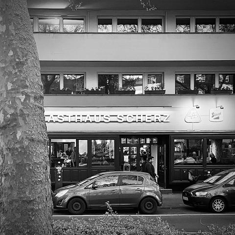 auf dem schwarz-weiß-foto ist die fassade eines gebäudes mit der aufschrift „gasthaus scherz“ zu sehen. davor stehen zwei autos auf der straße geparkt. ein großer baumstamm ragt links ins bild. daher ist von „gasthaus“ nur „asthaus“ zu lesen. das gasthaus hat große fenster, durch die man menschen im inneren beim essen oder sprechen erkennen kann. über dem schriftzug befinden sich wohnungen mit balkonen und blumenkästen. rechts an der fassade sind die logos von „mühlen kölsch“ und „stiegl“ zu sehen. die szene wirkt urban und belebt.