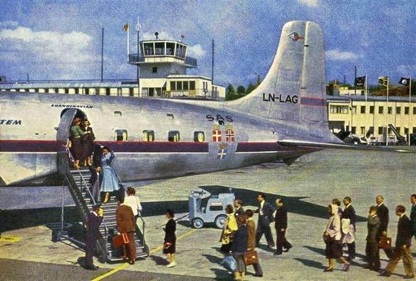 Only the tail of the airplane is seen in the center of the postcard. In the back is the tower of the airport. In the foreground are passengers queuing up to board.