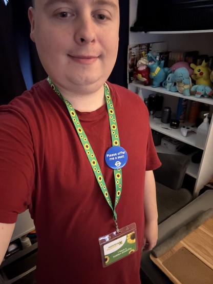 A white person is standing up. They are wearing a red t-shirt and a green Hidden Disabilities sunflower lanyard with “I have a hidden disability” on the green card attached to it. There is a Transport for London ‘Please Offer Me a Seat’ round blue badge with white letter pinned to the lanyard.