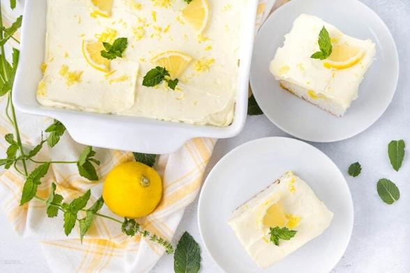 A lemon sheet cake topped with creamy frosting, lemon slices, and mint leaves. The cake is partially sliced in a white baking dish next to two plated servings adorned with similar garnishes. Fresh mint and a whole lemon are placed next to the dishes.