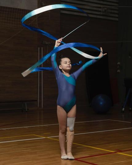 The image depicts a young girl engaged in rhythmic gymnastics. She is wearing a blue leotard with a gradient design and is performing with a blue ribbon. The girl is standing on a wooden gymnasium floor, and she appears to be in the middle of a routine, gracefully manipulating the ribbon with both hands. She is also wearing a prosthetic leg on her left side, which extends from her foot to above her knee. The background shows a typical indoor sports setting with a darkened area, possibly bleachers or a stage. The focus is on the gymnast's poised and concentrated expression as she executes her performance.