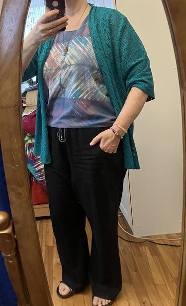 A woman in front of a mirror wearing a Shibori dyed cotton tank top on blues and purples , under a bright green shawl collar sweater with elbow length sleeves in an open weave fabric, and black linen drawstring pants with pockets.