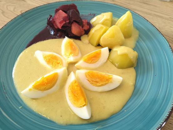 Plate of neatly arranged mustard eggs. Cooked beetroot with apple shavings in a jellied sauce of black currant and vinegar on the top left, cooked potatoes with splashes of mustard sauce on the top right. Bottom and center of the plate is occupied by 6 egg quarters arranged in the shape of a flower on a mirror of mustard sauce. // Hübsch angerichtete Senfeier auf einem blauen Teller. Oben links wurde die Rote Beete auf dem Teller arrangiert. Die Rote Beete ist verfeinert mit gehobelten Äpfelchen in einer Soße aus Johannisbeergelee, abgeschmeckt mit Apfelessig. Oben rechts sind gekochte Kartoffeln, die vorsichtig mit Senfsoße beträufelt wurden. Die untere Hälfte, sowie die MItte des Tellers nehmen die Eier ein. Es sind 6 Eiviertel, die in Blütenform auf einem Spiegel aus Senfsoße angeordnet wurden.