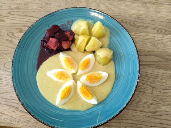 Plate of neatly arranged mustard eggs. Cooked beetroot with apple shavings in a jellied sauce of black currant and vinegar on the top left, cooked potatoes with splashes of mustard sauce on the top right. Bottom and center of the plate is occupied by 6 egg quarters arranged in the shape of a flower on a mirror of mustard sauce. // Hübsch angerichtete Senfeier auf einem blauen Teller. Oben links wurde die Rote Beete auf dem Teller arrangiert. Die Rote Beete ist verfeinert mit gehobelten Äpfelchen in einer Soße aus Johannisbeergelee, abgeschmeckt mit Apfelessig. Oben rechts sind gekochte Kartoffeln, die vorsichtig mit Senfsoße beträufelt wurden. Die untere Hälfte, sowie die MItte des Tellers nehmen die Eier ein. Es sind 6 Eiviertel, die in Blütenform auf einem Spiegel aus Senfsoße angeordnet wurden.