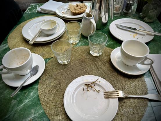 A table setting with several empty, white plates, bowls, coffee cups & glasses, all arranged on a textured placemat over a green marble table